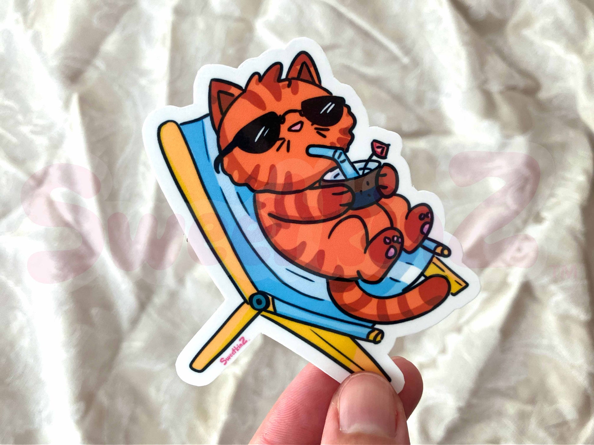 Sticker of a cat lounging on a beach chair with sunglasses and a drink, held against a textured white background.