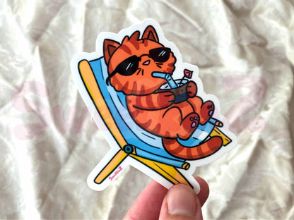 Sticker of a cat lounging on a beach chair with sunglasses and a drink, held against a textured white background.