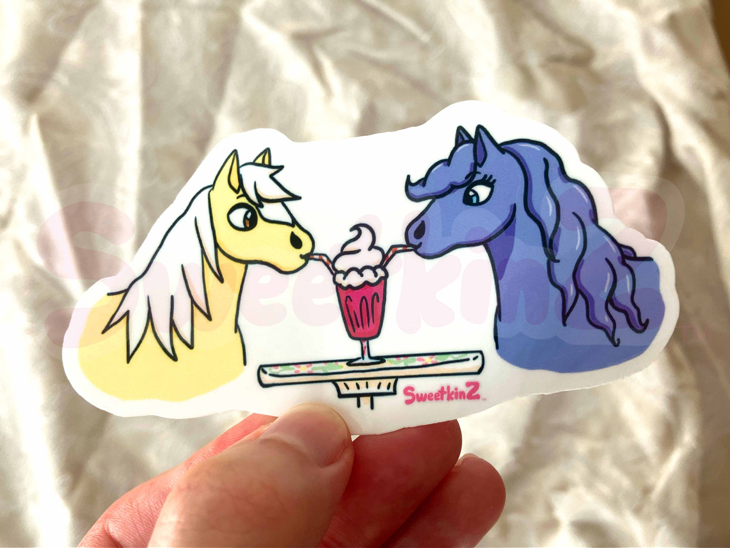 Sticker of two ponies sharing a milkshake, held by a hand against a neutral background